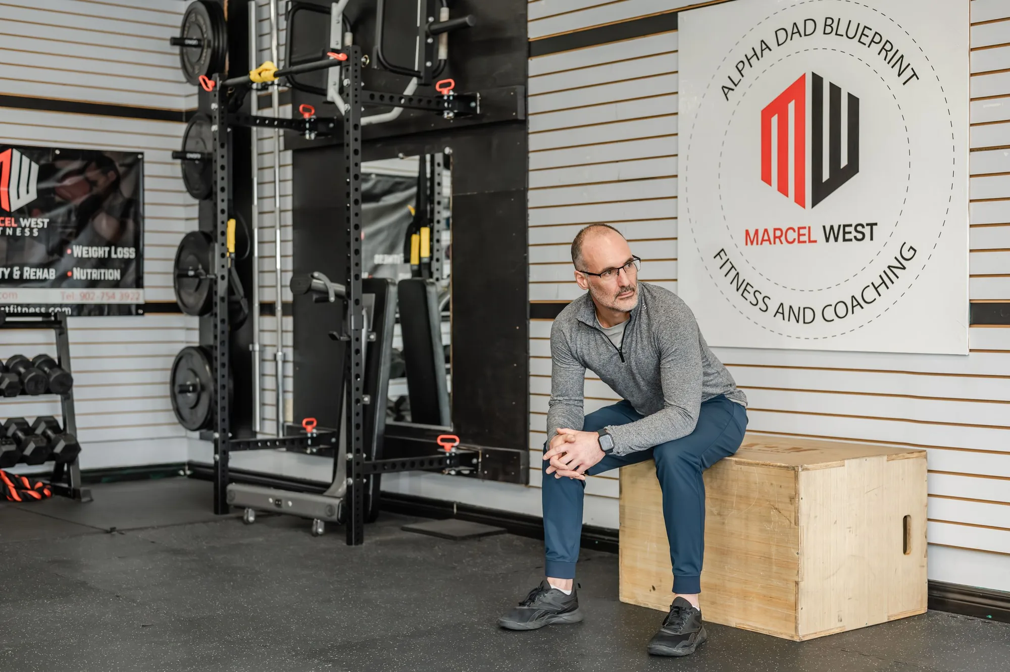 Marcel West seated on a plyo box in the MW Fitness and Coaching studio, beside the rig and the studio wall sign