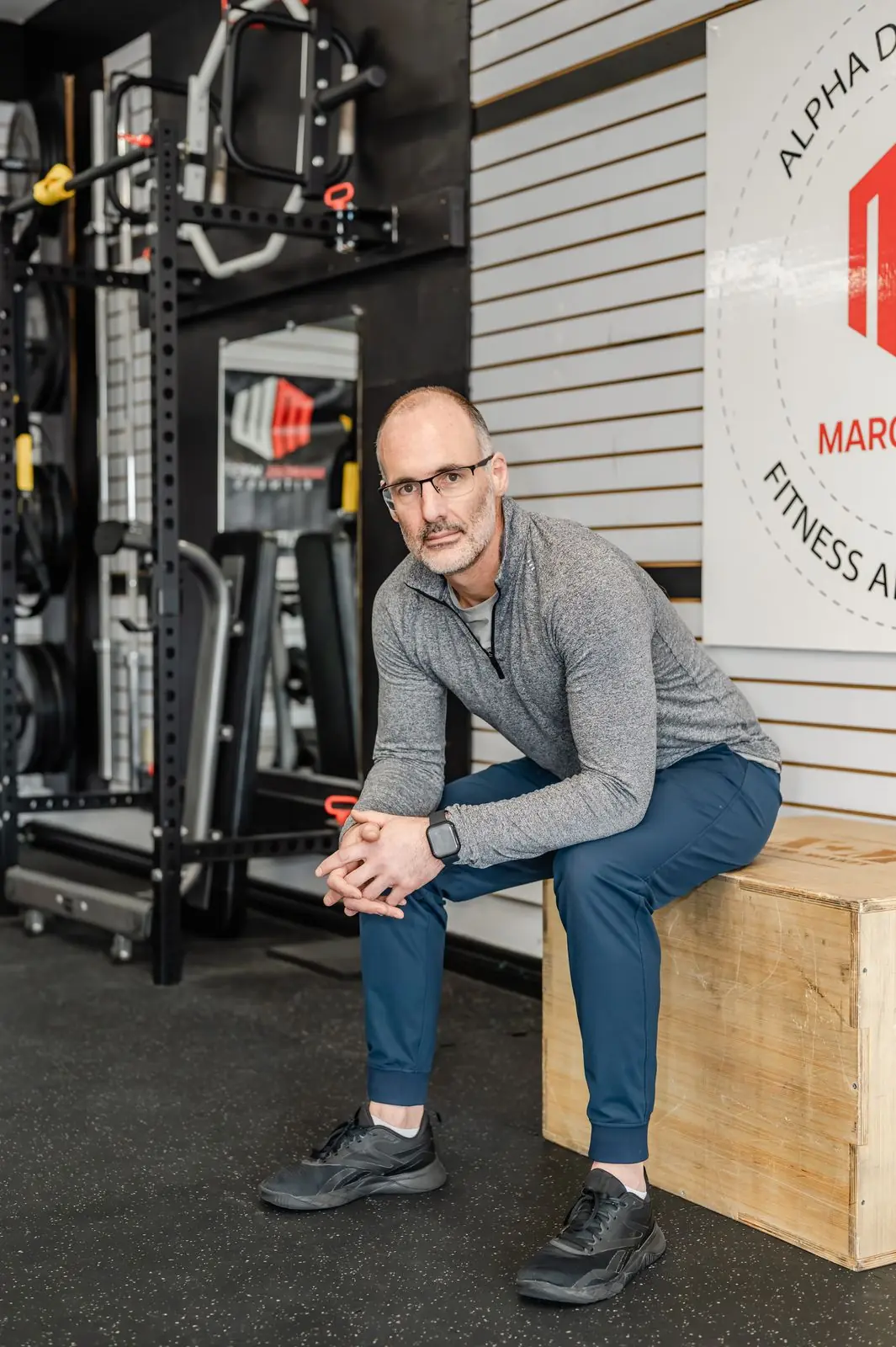 Marcel West seated on a plyo box beside the squat rig in the studio